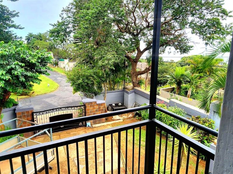 An elevated balcony view overlooking the front garden, driveway, and surrounding neighborhood greenery.