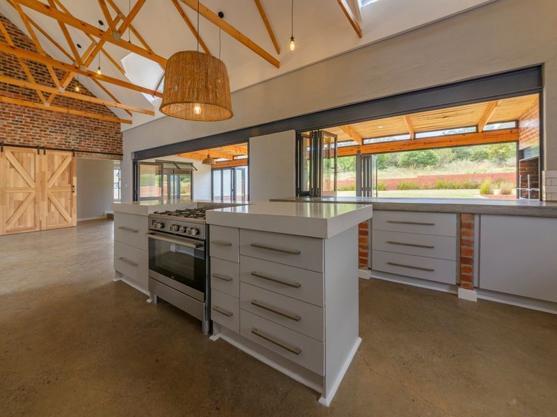 Spacious open-plan kitchen and living area featuring exposed timber trusses, brick accents, polished concrete floors, and seamless indoor-outdoor flow through large bi-fold doors.