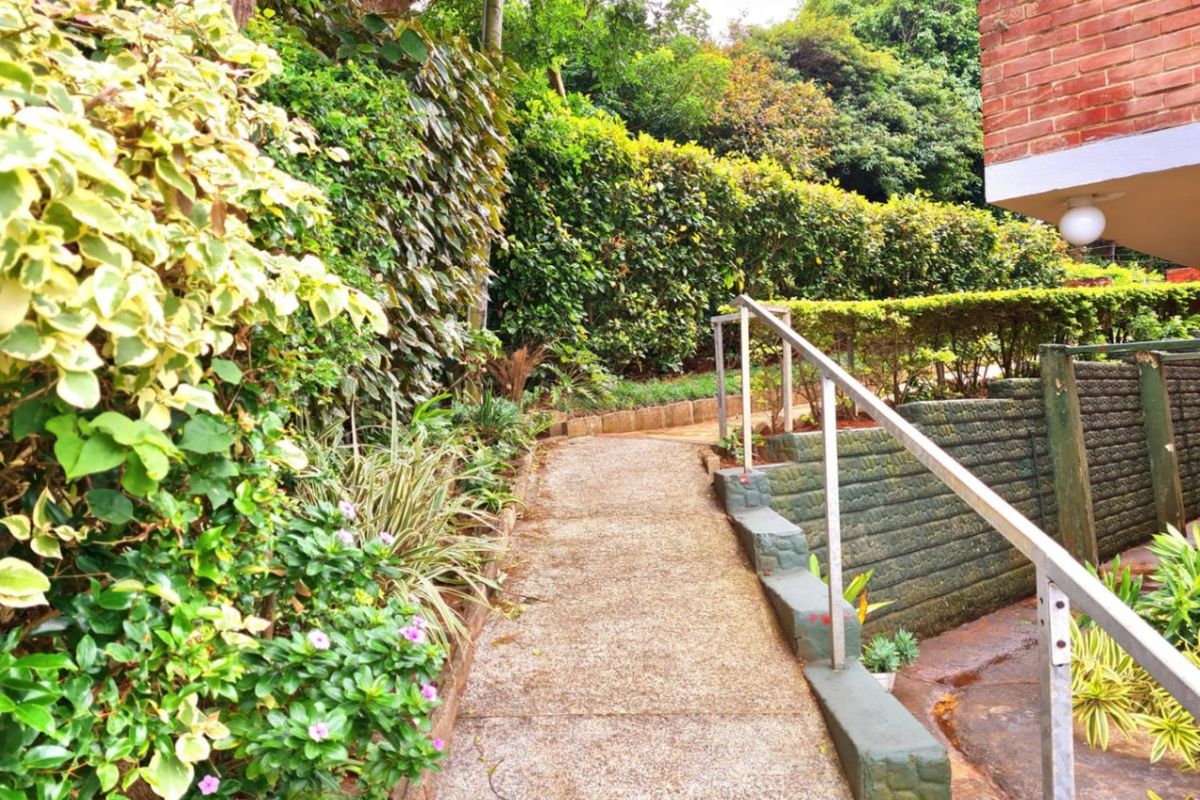 A paved garden pathway featuring a metal handrail and established landscaping, providing access along the side of the brick building structure.