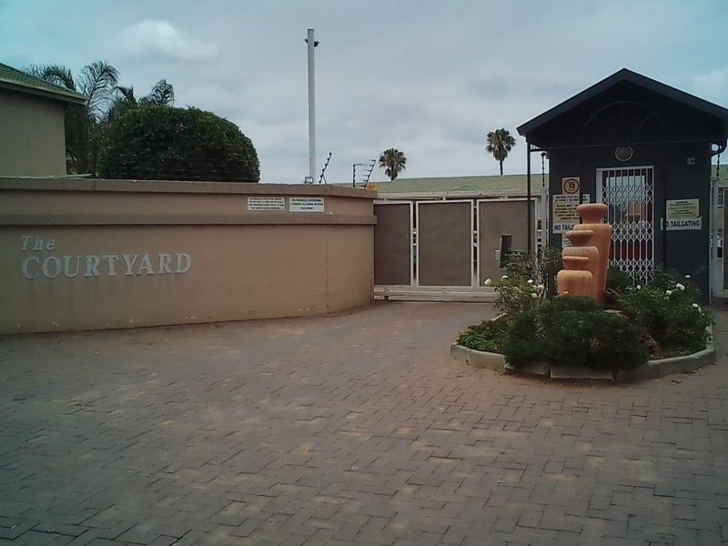 The entrance to The Courtyard apartment complex, featuring a paved driveway, security gate, and guard house with perimeter wall and electric fencing.