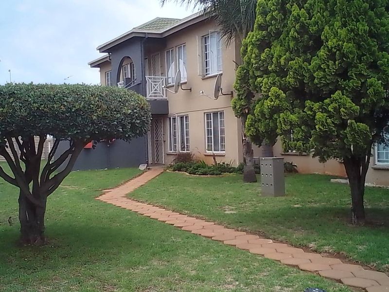Front exterior view of a multi-story apartment complex featuring a paved walkway, established garden, and security-gated entrances.