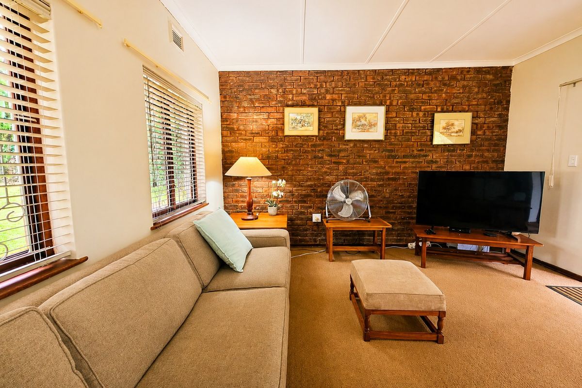 A comfortable living room featuring an exposed brick accent wall, neutral carpeting, and large windows with venetian blinds providing natural light.