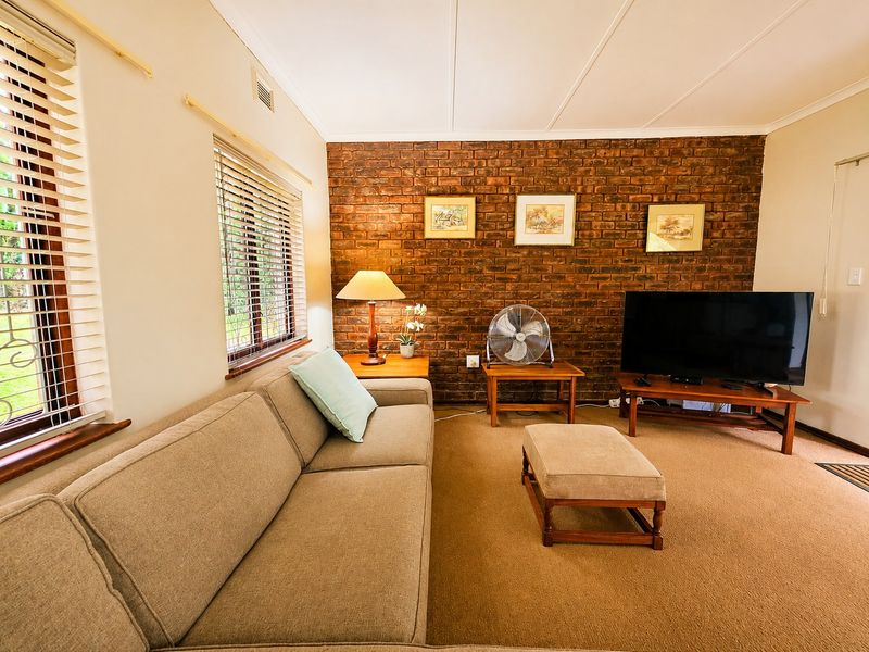 A comfortable living room featuring an exposed brick accent wall, neutral carpeting, and large windows with venetian blinds providing natural light.