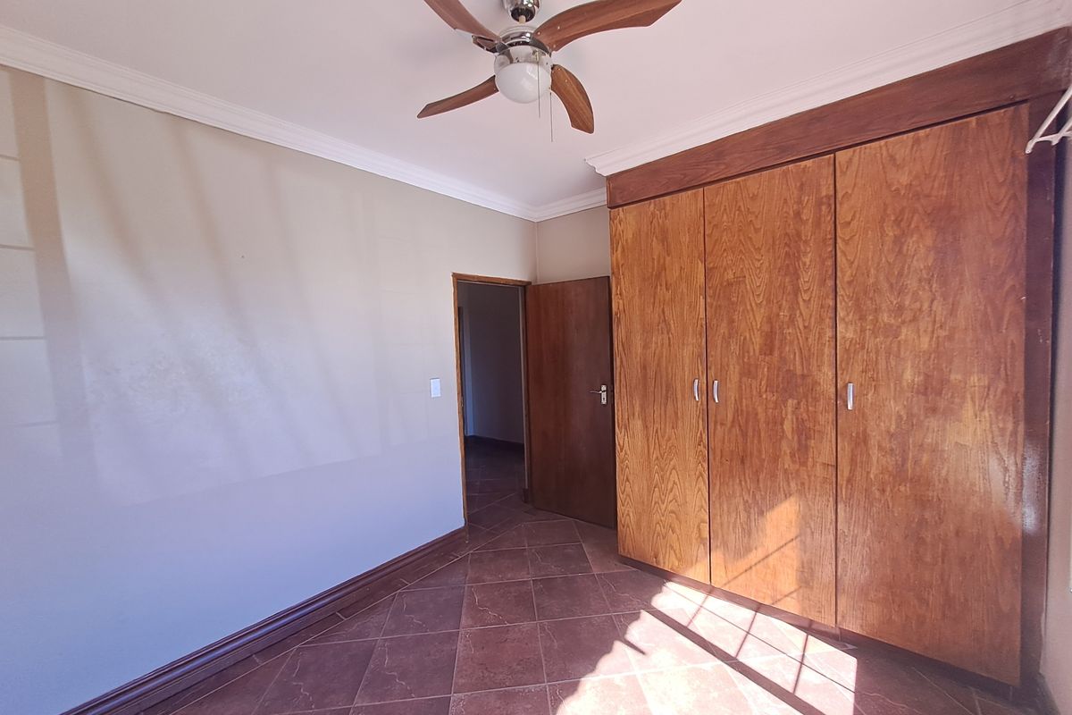 A spacious bedroom featuring built-in wooden cupboards, tiled flooring, and a ceiling fan, offering a clean and functional space.