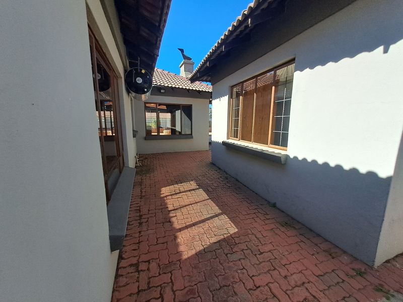 A paved exterior walkway between two sections of the house, featuring tiled roofing, timber-framed windows, and an external geyser unit.