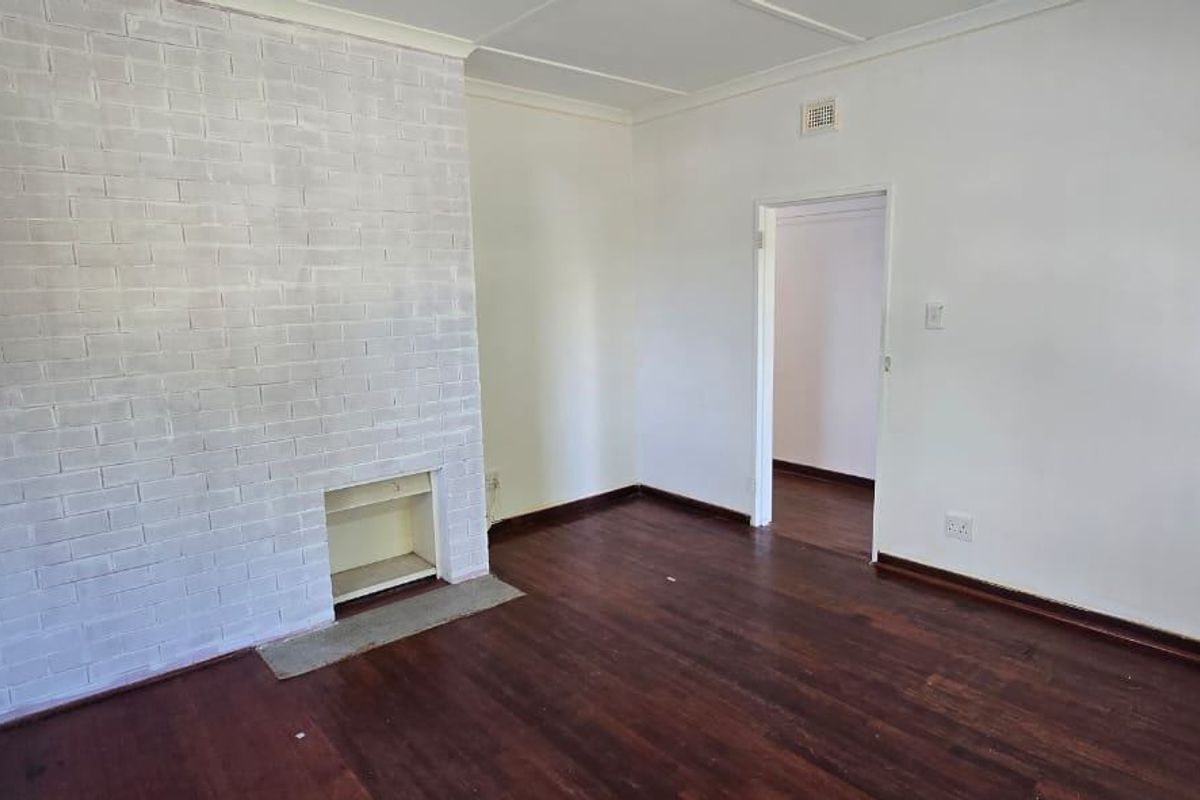 An empty living room featuring original hardwood flooring and a painted brick fireplace, offering a blank canvas for interior design.