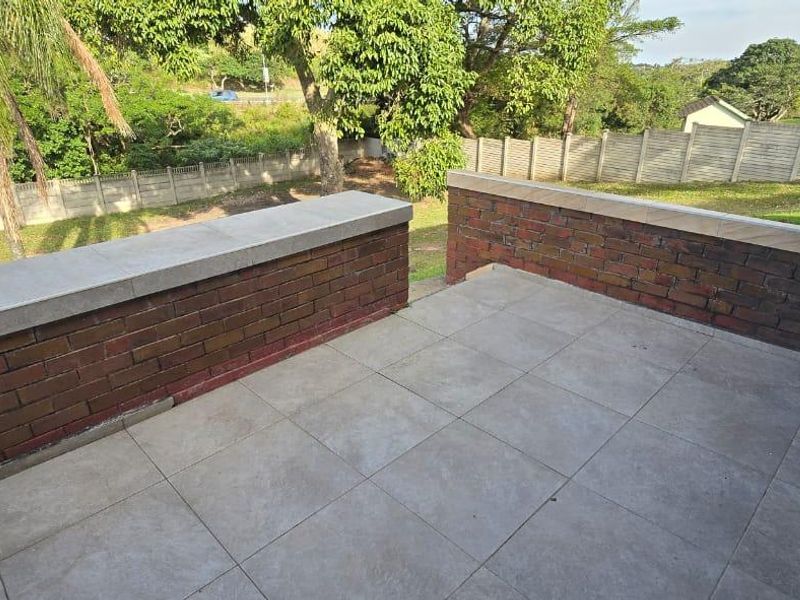 An open-air balcony featuring tiled flooring and a brick balustrade with stone capping, overlooking the surrounding garden area.