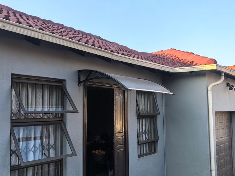 Exterior view of the house entrance featuring a tiled roof, grey painted facade, security windows, and a small covered porch area.