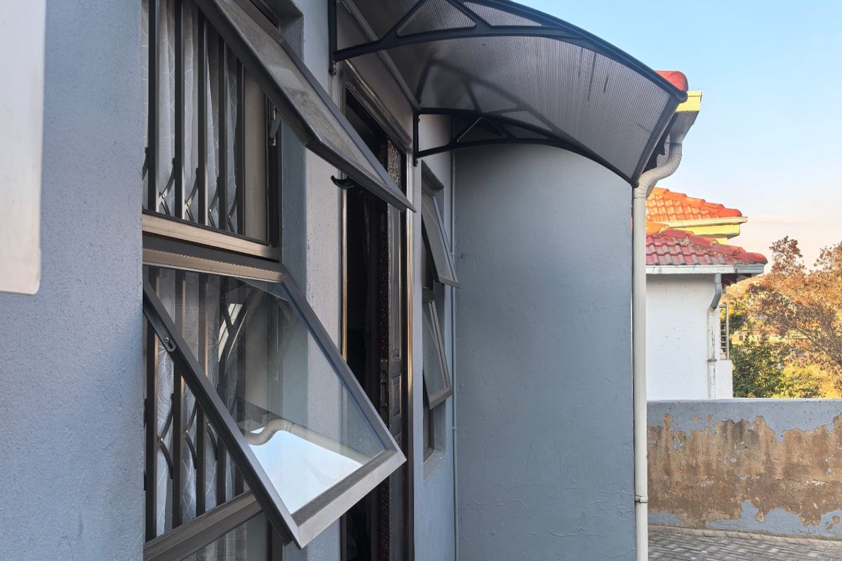 Exterior view of the side entrance featuring a modern polycarbonate door awning, top-hung windows with security bars, and tiled step access.