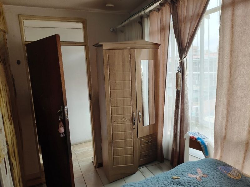 A bedroom featuring tiled flooring, a wooden door, and a large window with curtains, showing signs of wear and requiring cosmetic updates.