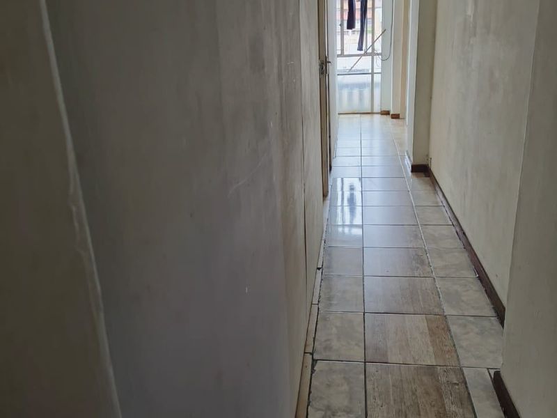 A narrow, empty hallway featuring tiled flooring and plain walls, leading towards a rear exit or balcony area.