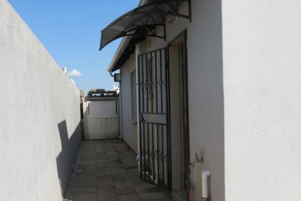 A side exterior walkway featuring a paved path, security gate, protective awning, and boundary wall, providing functional access to the property.