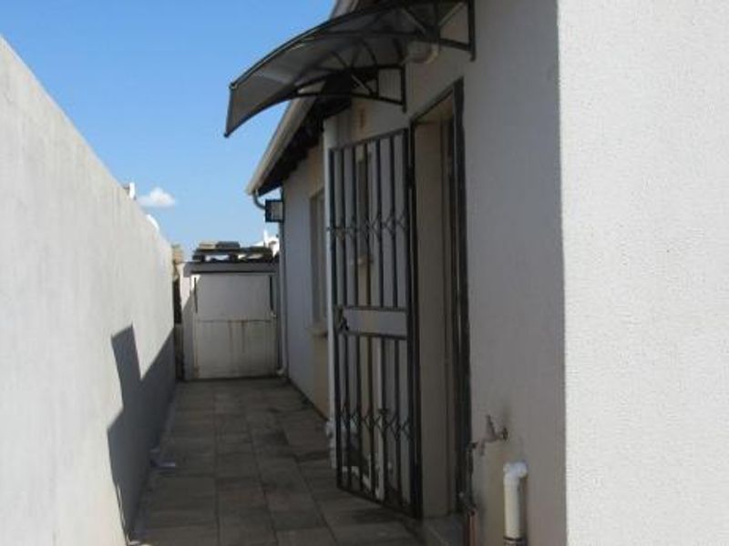 A side exterior walkway featuring a paved path, security gate, protective awning, and boundary wall, providing functional access to the property.