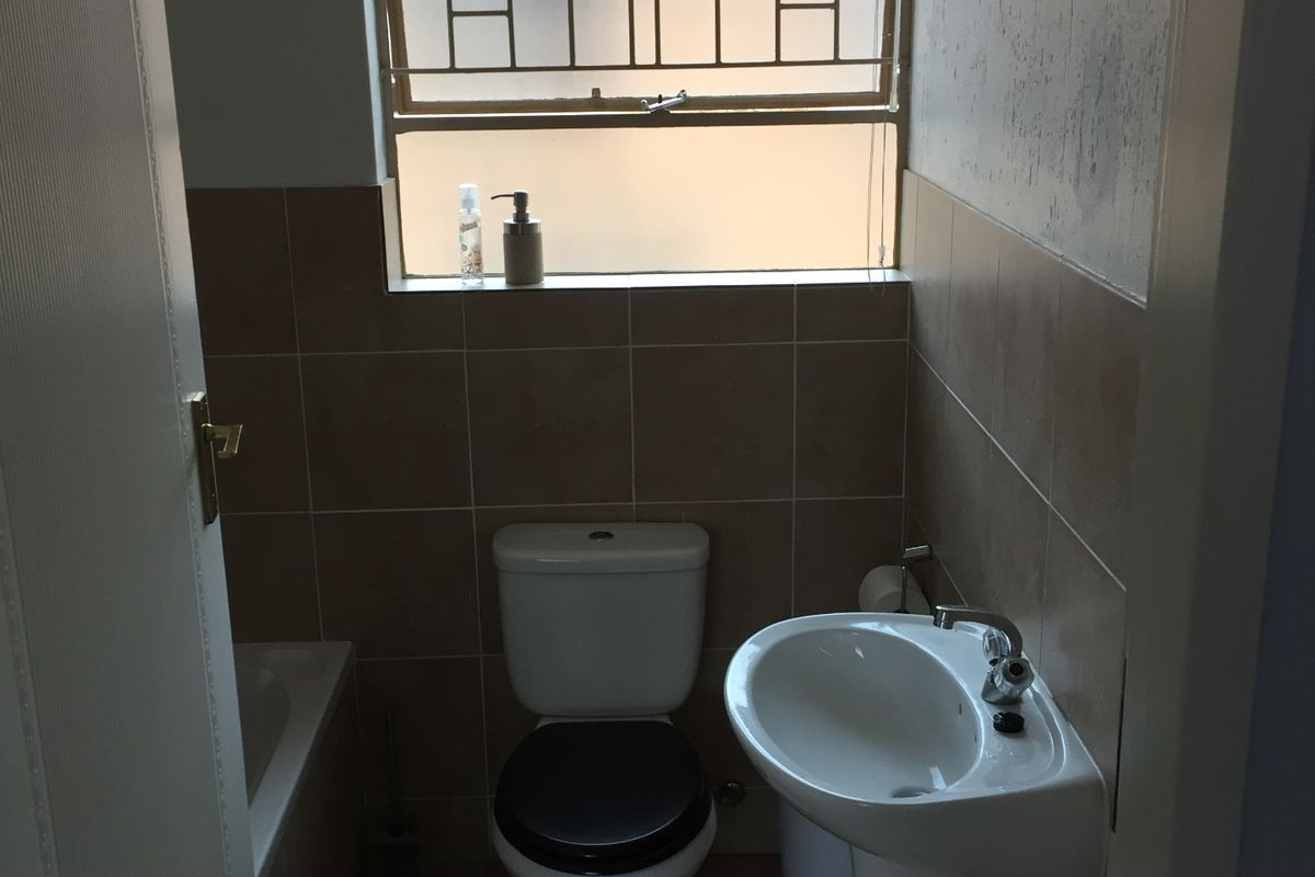 A compact bathroom featuring a pedestal basin, toilet, and bathtub with tiled finishes and a window with security bars.
