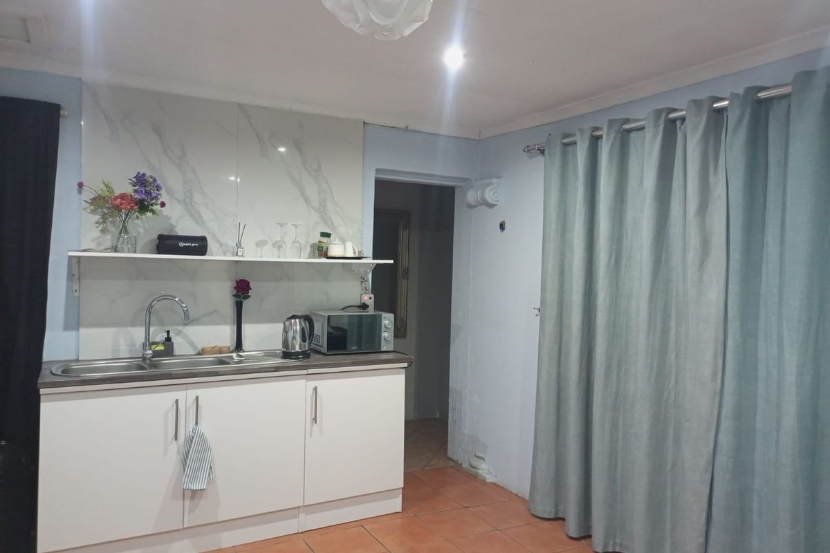 A compact kitchen area featuring white cabinetry, a marble-look splashback, stainless steel sink, and tiled flooring, requiring some wall maintenance.