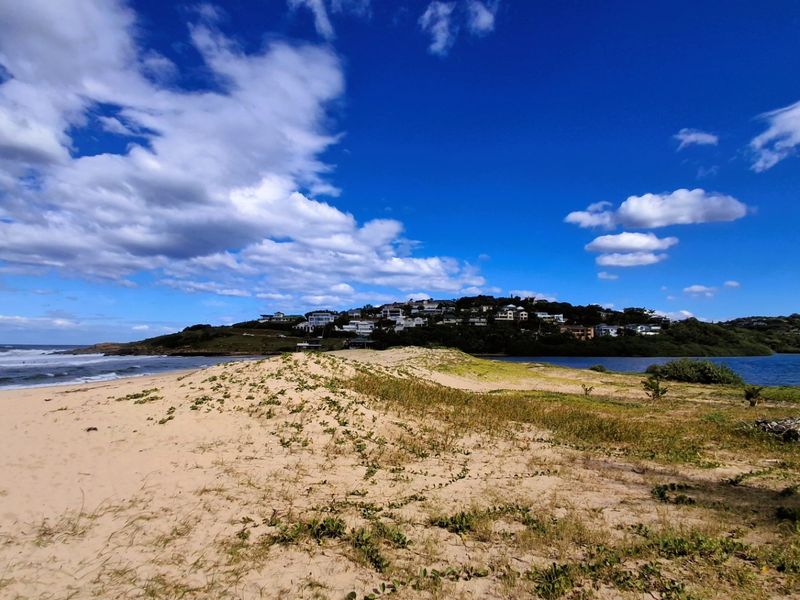 A scenic view of the coastal landscape featuring sand dunes, ocean waves, and an estuary with residential properties visible in the distance.