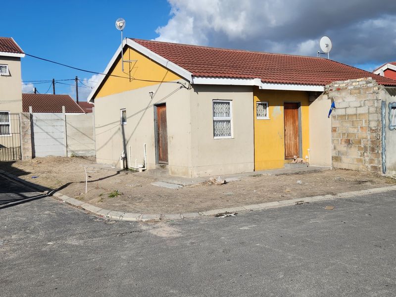 A detached single-story house featuring a tiled roof, yellow and beige exterior walls, and a corner lot position with an unpaved yard.