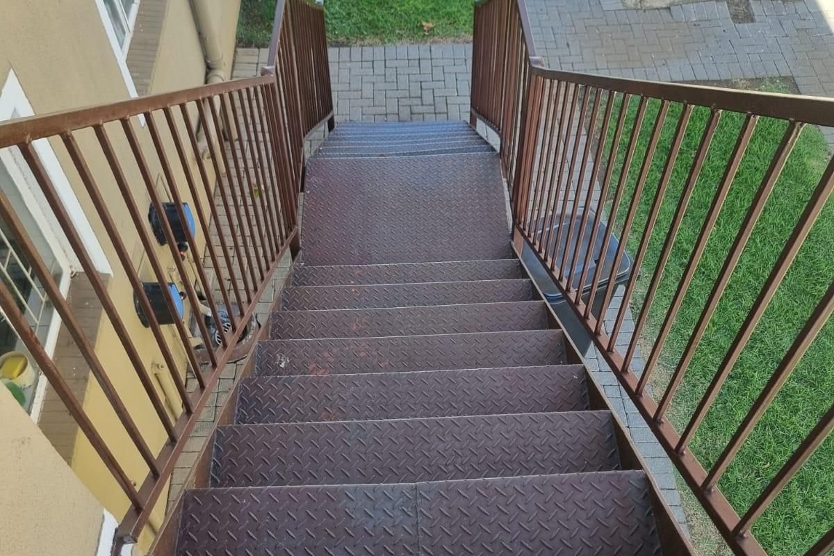 Exterior metal staircase leading down to a paved walkway and garden area, showing signs of surface rust and wear.