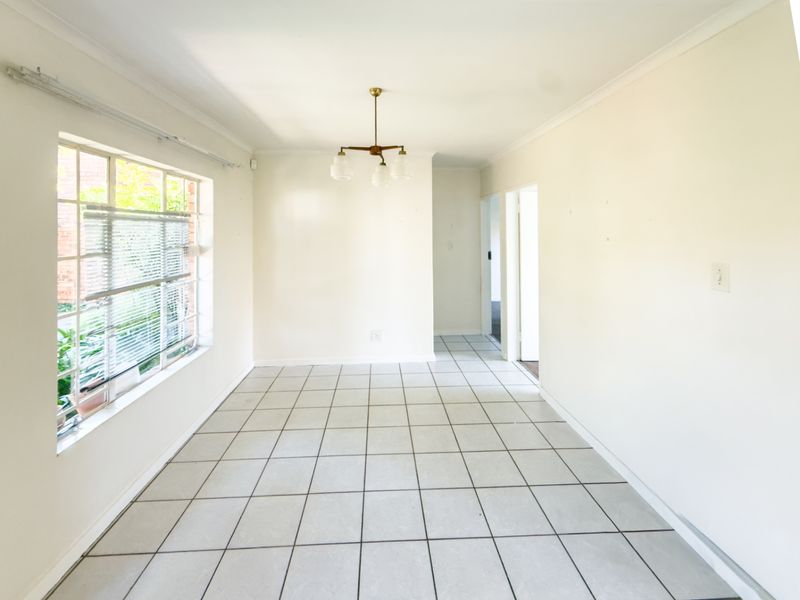 A bright, empty dining room featuring neutral tiled flooring, a central light fixture, and a large window providing natural light.
