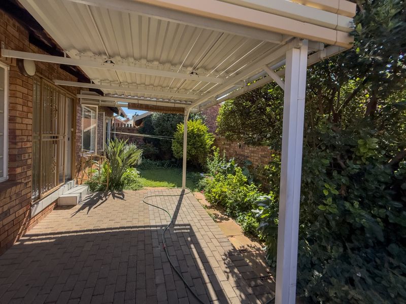 A covered brick-paved patio area overlooking a private garden, featuring a metal roof structure and direct access to the home.