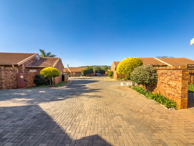 A view of the paved driveway and entrance area within a townhouse complex, featuring face brick construction and established landscaping.