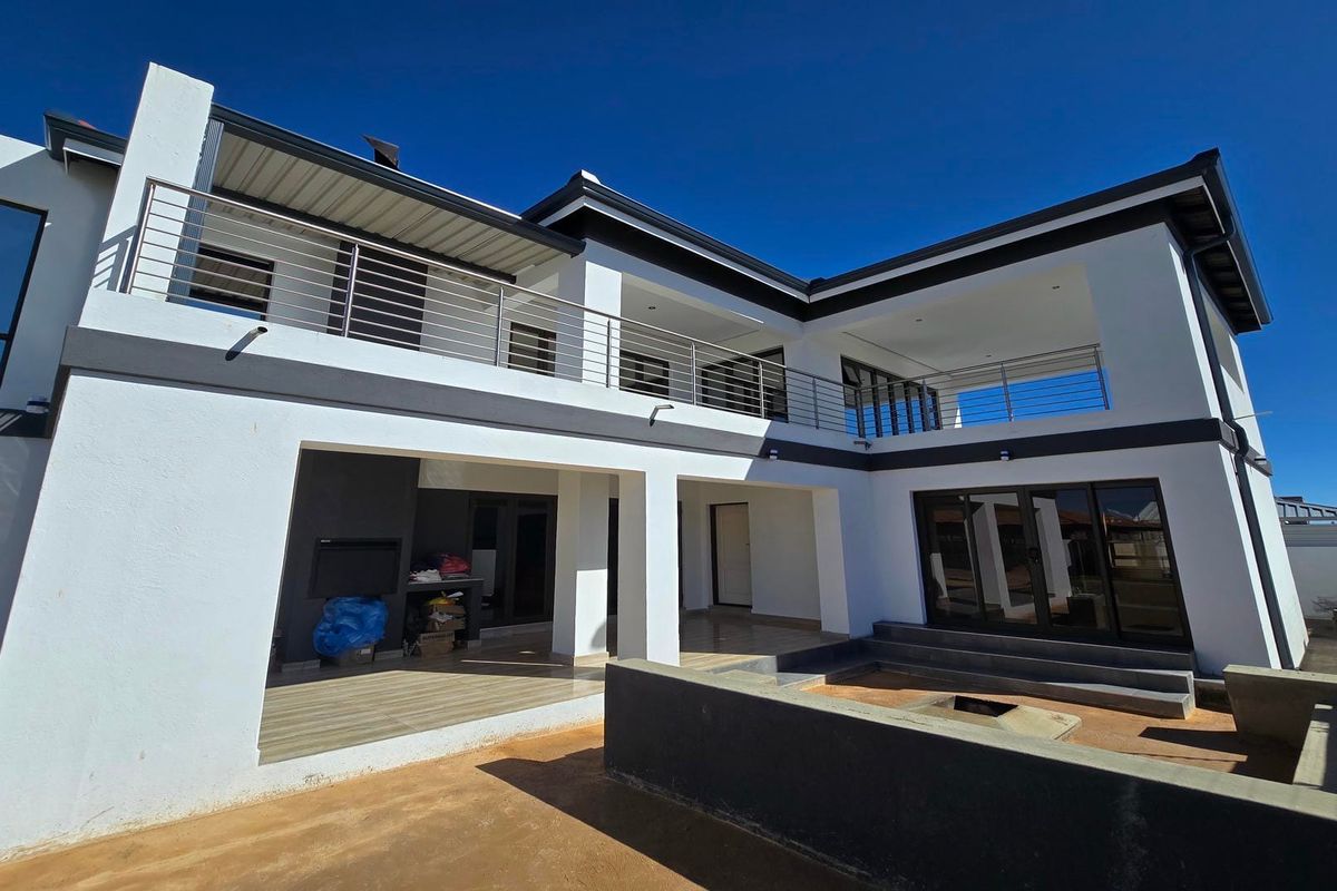 Modern double-story rear elevation featuring a spacious covered patio, built-in braai, upper-level balcony with stainless steel balustrades, and a sunken fire pit area.