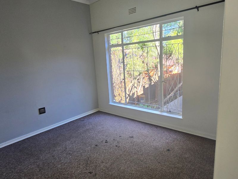 An empty bedroom featuring grey walls, carpet flooring, and a large window with security bars, offering natural light and a view of the exterior.