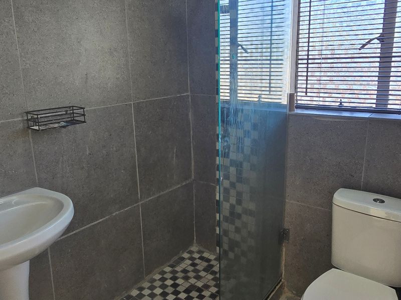 A functional bathroom featuring a pedestal basin, a glass-enclosed shower with mosaic floor tiles, and a standard toilet with grey wall tiling.