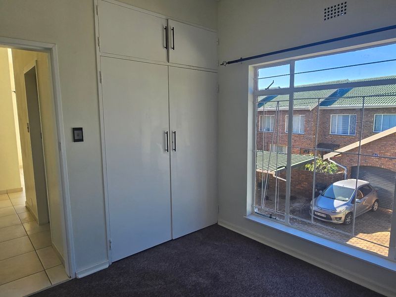 A bright, empty bedroom featuring built-in cupboards, carpeted flooring, and a large window with security bars overlooking the exterior.