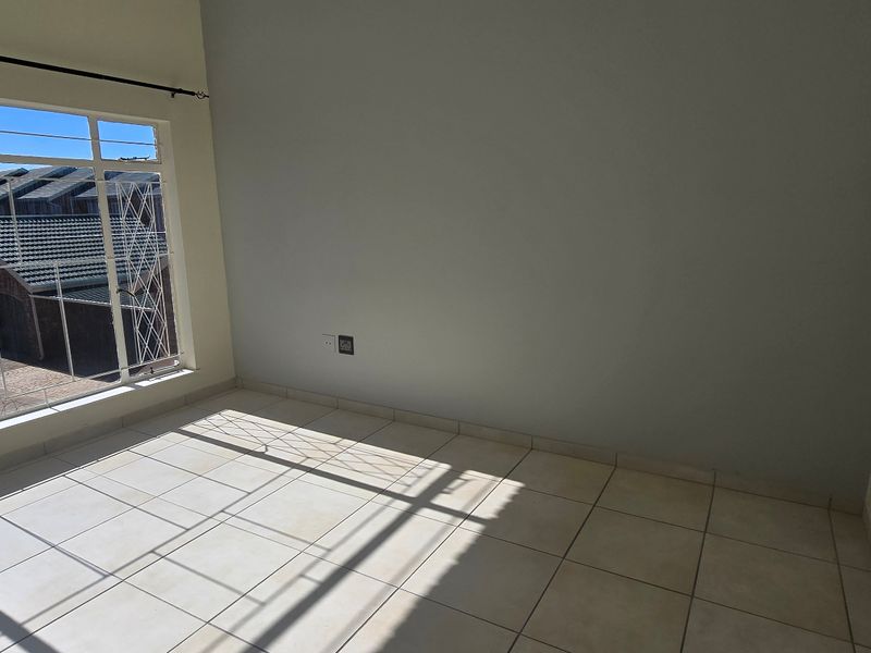 A bright, empty bedroom featuring neutral tiled flooring, a large window with security bars, and clean wall finishes.