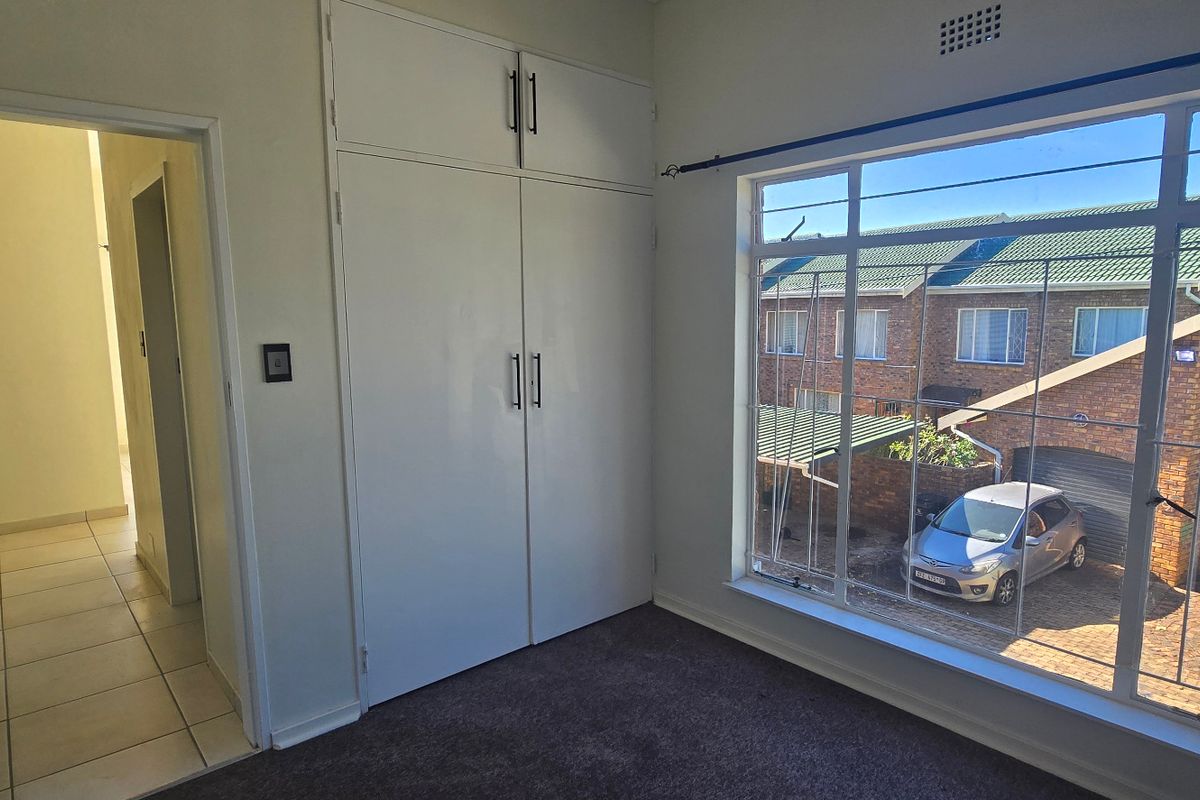 A bright, empty bedroom featuring built-in cupboards, carpeted flooring, and a large window with security bars overlooking the exterior.