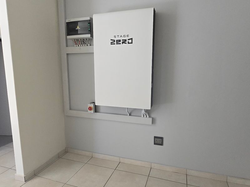A clean utility space featuring a wall-mounted solar battery storage system and electrical distribution board, finished with neutral tiled flooring.