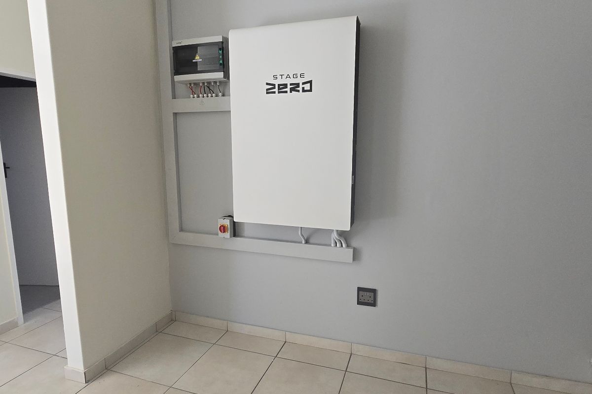A clean utility space featuring a wall-mounted solar battery storage system and electrical distribution board, finished with neutral tiled flooring.