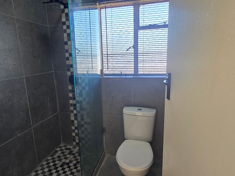 A modern bathroom featuring grey wall tiles, a glass-enclosed shower, a pedestal basin, and a standard toilet with natural light from the window.