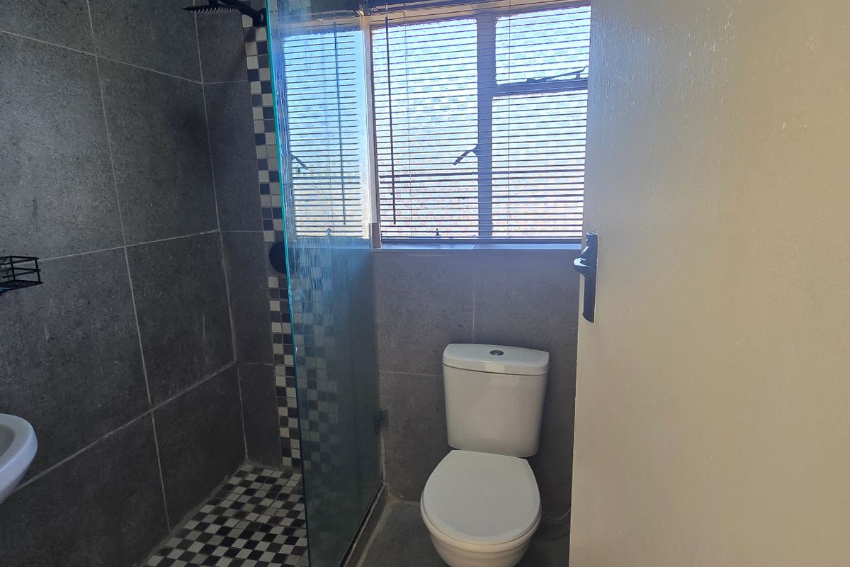 A modern bathroom featuring grey wall tiles, a glass-enclosed shower, a pedestal basin, and a standard toilet with natural light from the window.