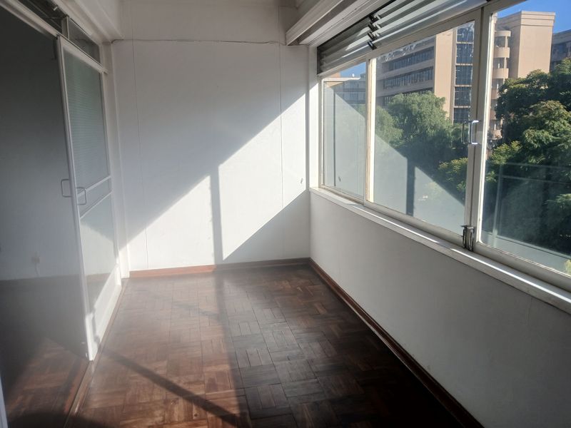 A sunlit enclosed balcony featuring classic parquet flooring and large windows, offering a versatile space for a home office or relaxation area.