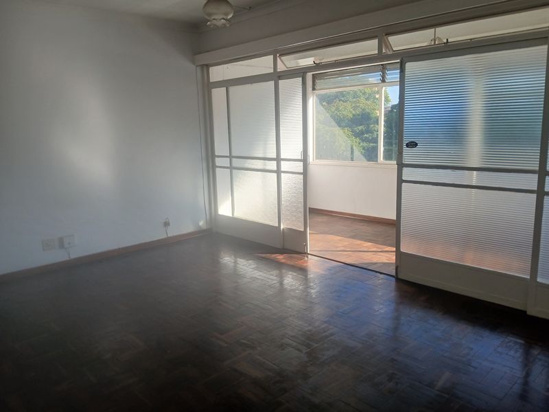A bright living room featuring original parquet flooring and large sliding glass doors that open onto an enclosed balcony area.