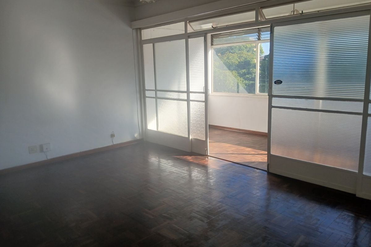 A bright living room featuring original parquet flooring and large sliding glass doors that open onto an enclosed balcony area.