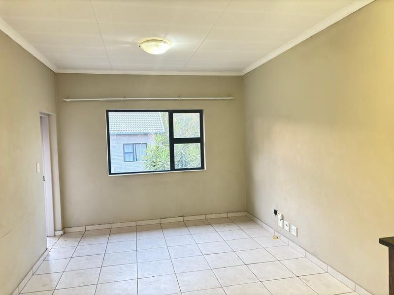 A bright, empty living area featuring neutral wall tones, tiled flooring, and a large aluminium-framed window providing natural light.