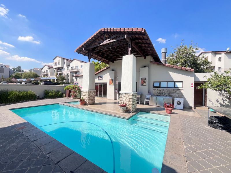 A well-maintained communal pool area featuring a covered patio with stone-clad pillars, tiled roofing, and paved surroundings within an apartment complex.