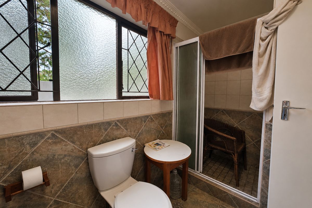 A dated bathroom featuring a toilet, shower enclosure, and tiled finishes, with a large window providing natural light.