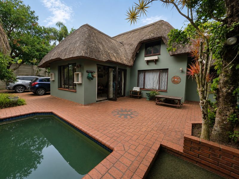 Rear view of a thatched-roof house featuring a swimming pool, brick-paved patio area, and established garden surroundings.