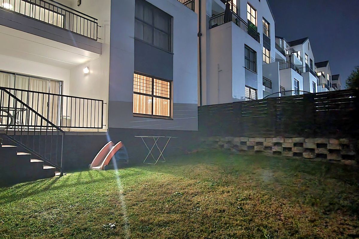 Night view of the rear exterior of the apartment building, featuring a private garden area, retaining wall, and a balcony with metal railings.