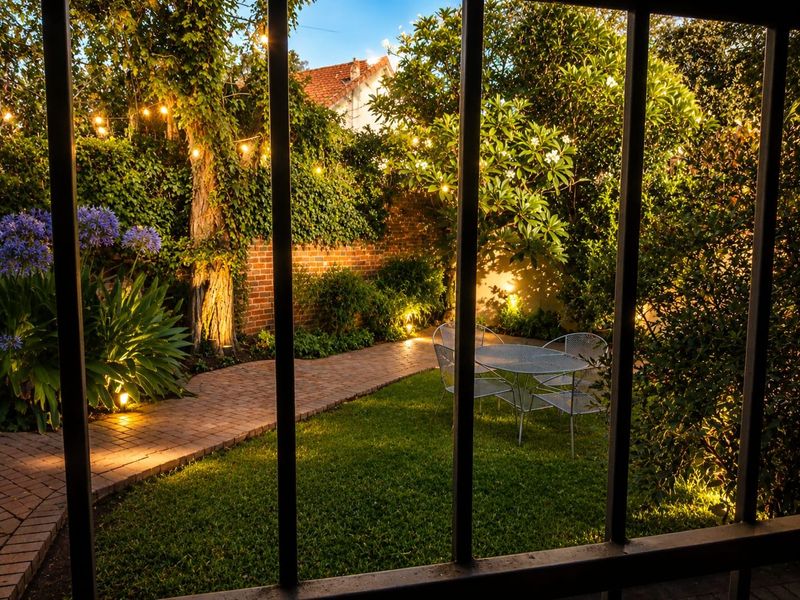 A beautifully landscaped private garden featuring a brick paved pathway, mature greenery, and ambient integrated lighting, viewed through a framed opening.