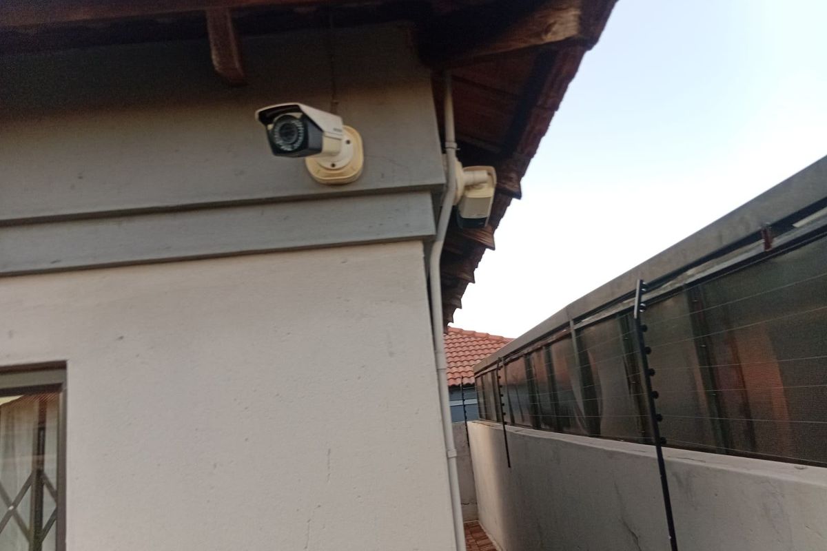 Exterior side passage featuring mounted security cameras and perimeter electric fencing along the boundary wall.
