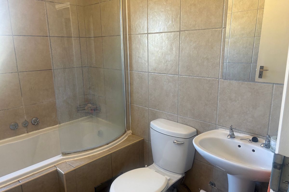 A functional bathroom featuring a bathtub with a curved glass shower screen, pedestal basin, and toilet, finished with neutral wall tiling.