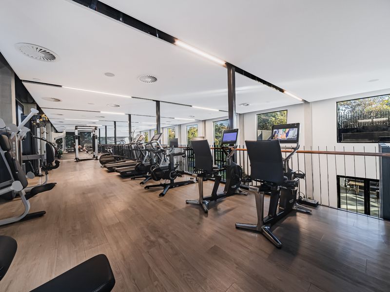 A modern, well-lit gym facility featuring timber flooring, a metal balustrade, and large windows providing natural light and views.