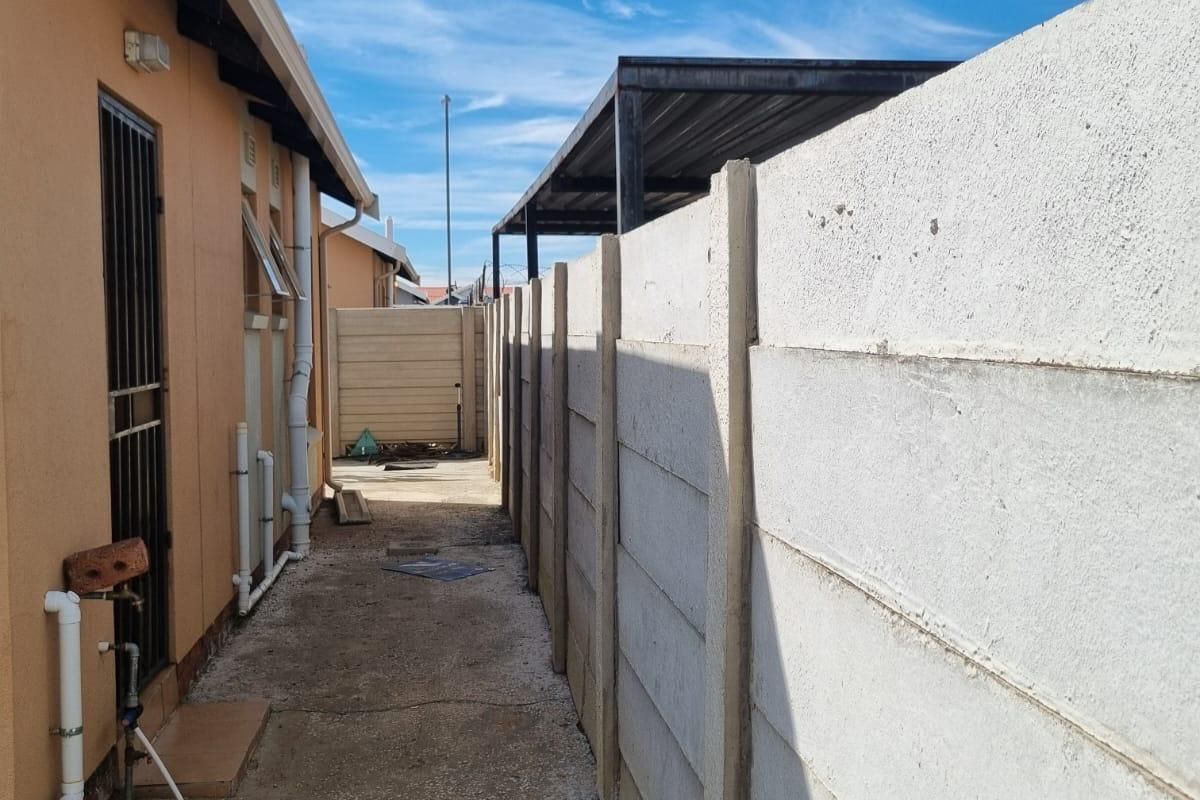 A narrow side passage of the property featuring a precast concrete boundary wall, external plumbing fixtures, and a metal carport structure at the rear.