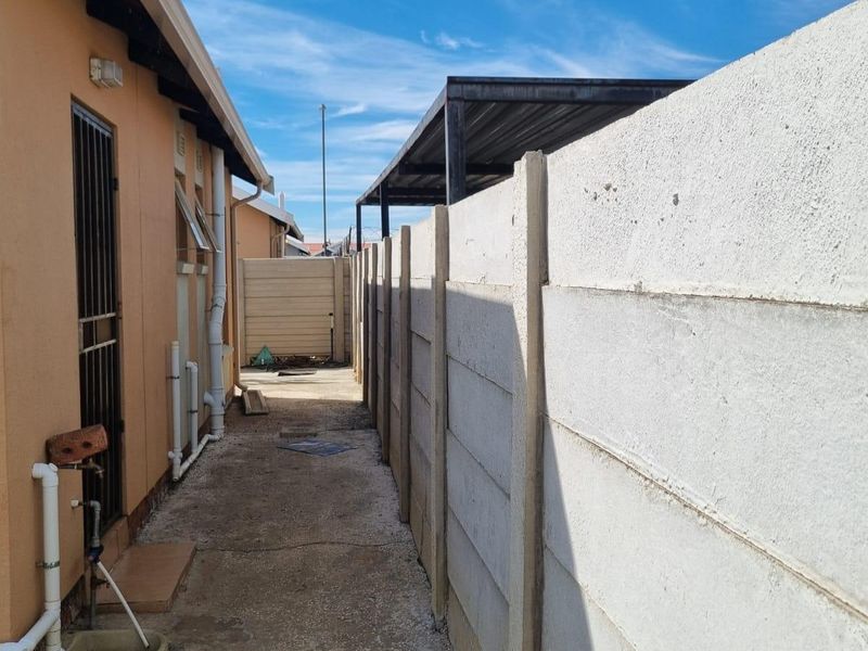 A narrow side passage of the property featuring a precast concrete boundary wall, external plumbing fixtures, and a metal carport structure at the rear.