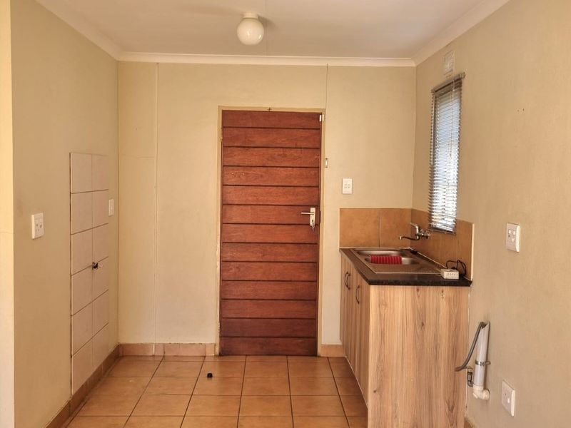 A compact, empty kitchen area featuring tiled flooring, built-in cabinetry with a stainless steel sink, and a wooden door leading to the exterior.
